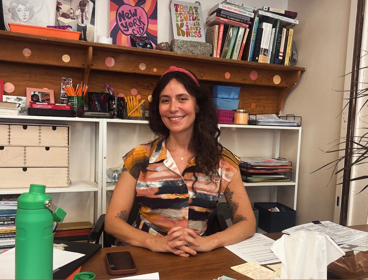 English teacher Ms. Walsh sits at her desk, sharing a welcoming smile with her students.